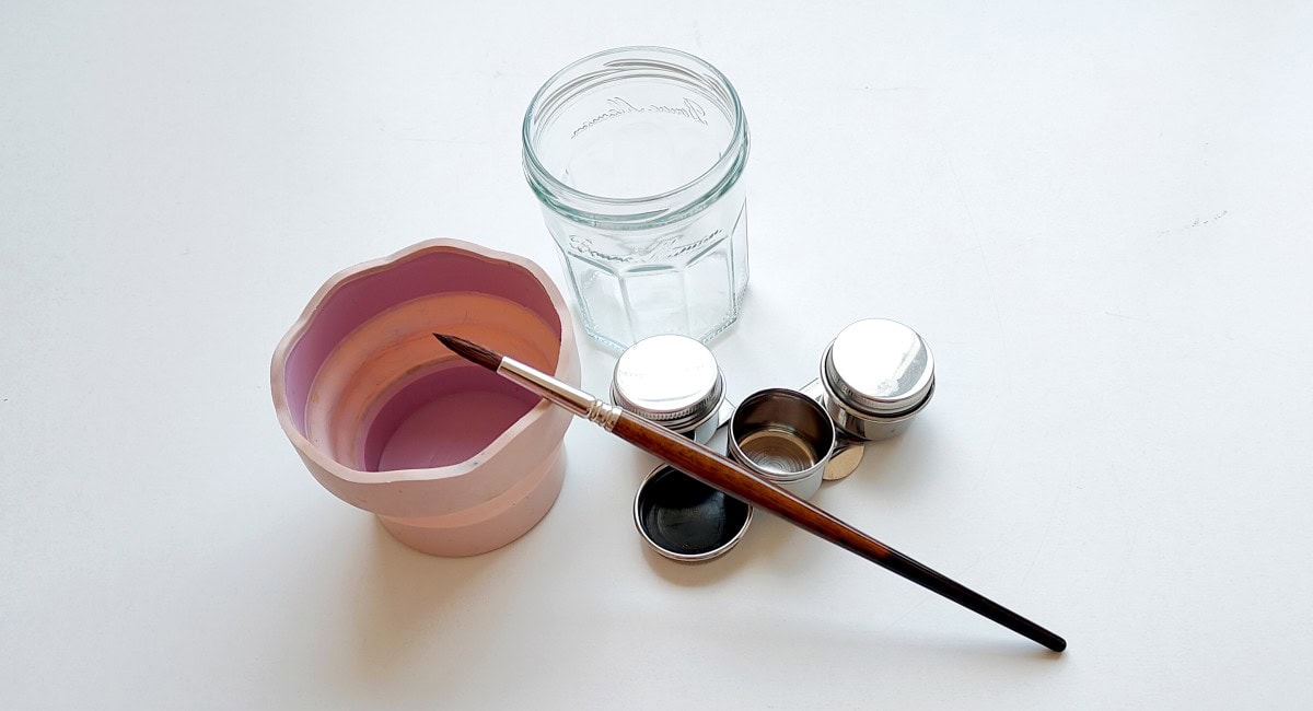 Varios vasos de agua para pintar con acuarela junto a pinceles y paleta.